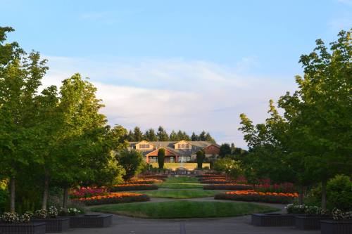 A landscaped garden with colorful flower beds, trees lining the path, and a house in the background under a blue sky.