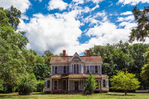 An old, abandoned house with a porch, surrounded by trees and under a partly cloudy sky.