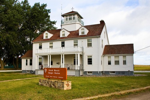 Historic white building with a tower, labeled "Grand Marais Ranger Station," surrounded by greenery and a cloudy sky.