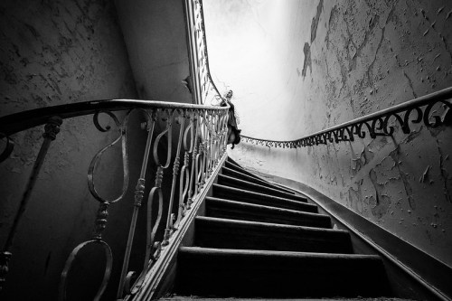 A winding staircase with a vintage railing, leading upward in a dimly lit, peeling wall environment.