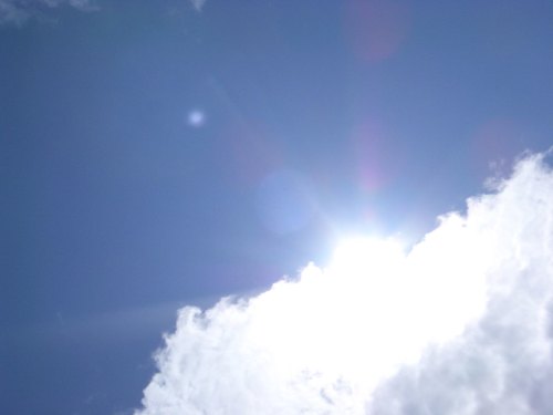 Bright sun shining in a clear blue sky, partially obscured by fluffy white clouds.