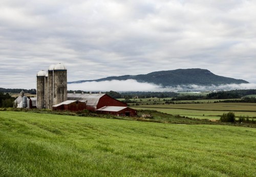 A scenic farm with silos and a red barn, set against a backdrop of rolling hills and misty mountains under a cloudy sky.