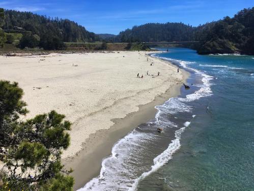 A sandy beach with people enjoying the sun, surrounded by lush green hills and a calm blue river.