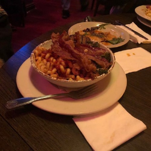 A bowl of macaroni topped with crispy bacon, served on a plate with a fork and napkin.