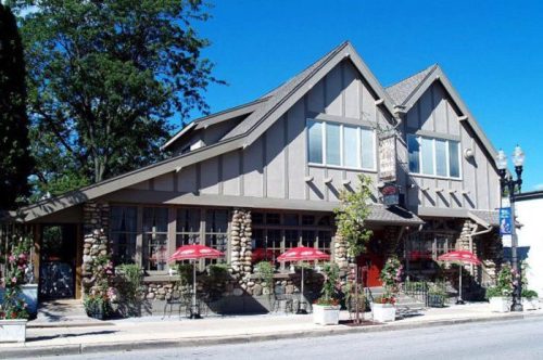A charming restaurant with a stone facade, large windows, and outdoor seating under red umbrellas on a sunny day.