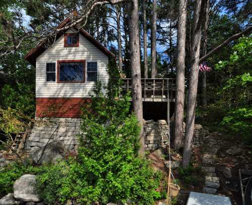 A small house nestled among trees, featuring a deck and surrounded by stone landscaping and greenery.