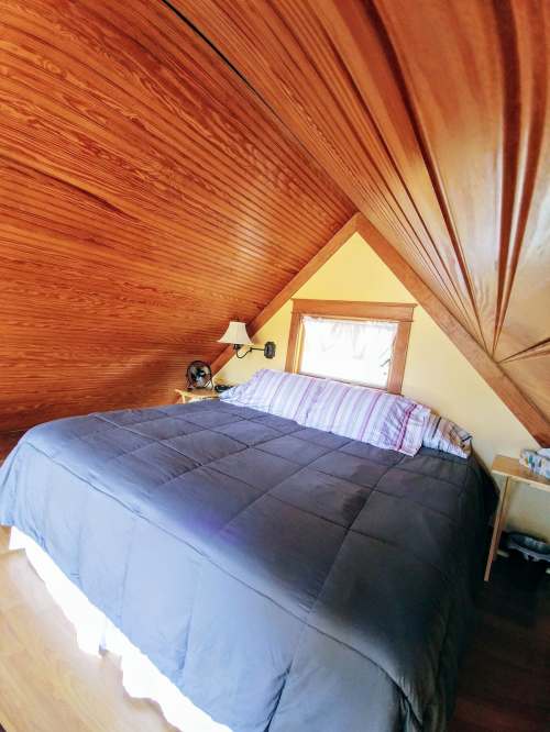 Cozy attic bedroom with a sloped wooden ceiling, a large bed, and a small nightstand by the window.