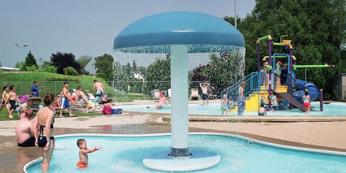 A water fountain in a pool area with children playing, adults relaxing, and a playground in the background.
