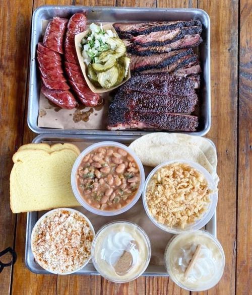 A metal tray with smoked sausage, brisket, pickles, beans, bread, and desserts, served with tortillas.
