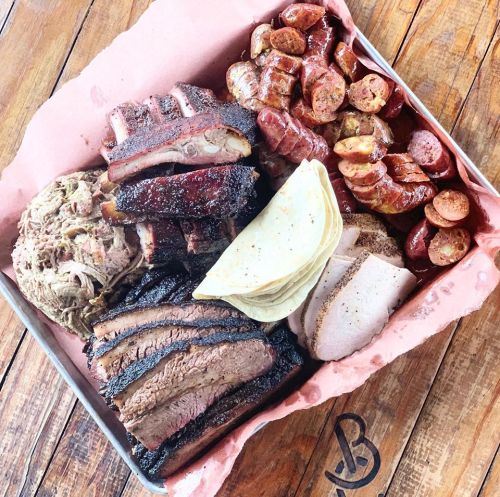 A platter of assorted barbecue meats including brisket, ribs, sausage, pulled pork, and tortillas on a wooden table.