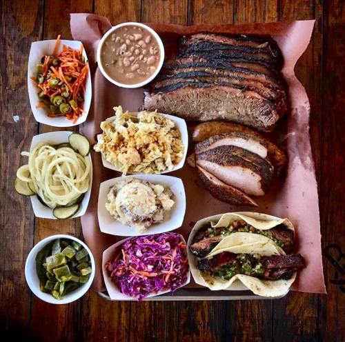 A platter of barbecue featuring brisket, sausage, tacos, and various sides like coleslaw, beans, and pickles.