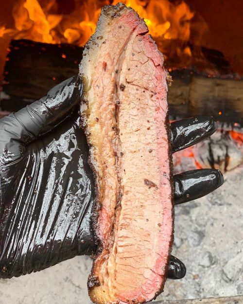 A hand in a black glove holds a slice of smoked brisket, with flames and logs in the background.