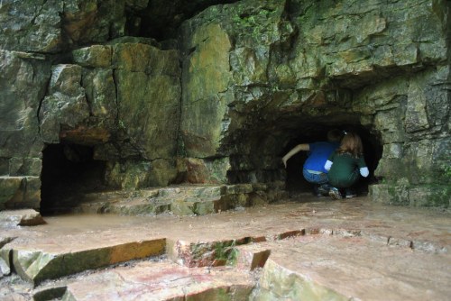 Two people exploring a rocky cave entrance, surrounded by moss-covered stone walls.