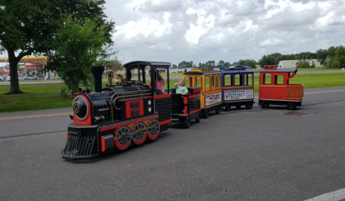 A small, colorful train with children riding on it, set against a cloudy sky and grassy area.