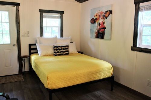 Cozy bedroom with a yellow bedspread, white pillows, and a colorful cow painting on the wall. Natural light from windows.