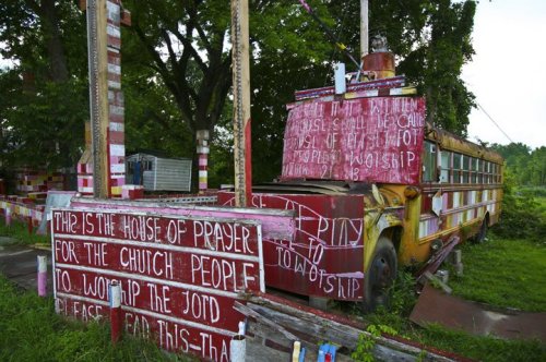 A colorful, graffiti-covered school bus in an overgrown area, with messages about prayer and worship painted on it.