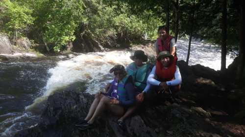 Four people sit by a river, surrounded by trees, enjoying a sunny day outdoors. Water flows rapidly in the background.
