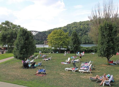 A scenic park by a river with people relaxing on lounge chairs and a bridge in the background.