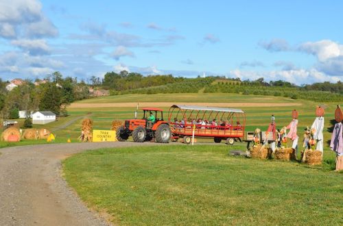 A red tractor pulls a hayride through a scenic farm with hay bales and scarecrows under a blue sky.