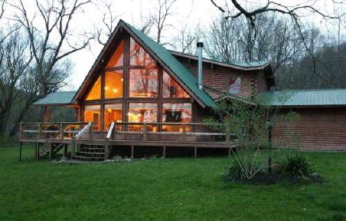 A wooden cabin with large glass windows, surrounded by trees and a grassy area, illuminated at dusk.