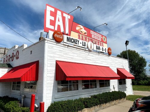 A barbecue restaurant with a red awning and a large "EAT" sign on the roof, featuring a clear blue sky in the background.