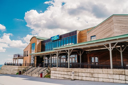 A modern wooden building with large windows, set against a blue sky with fluffy clouds, near a waterfront.