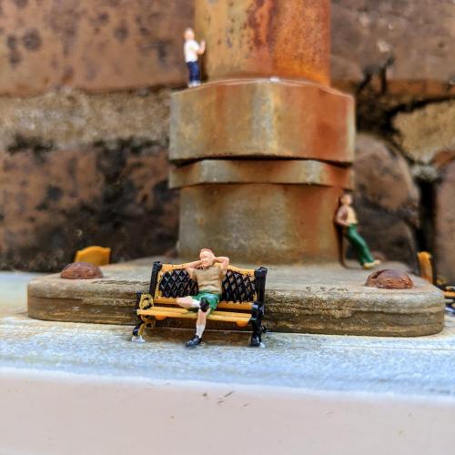 Miniature figures on a bench and a pipe, depicting a playful urban scene with a relaxed figure and another climbing.