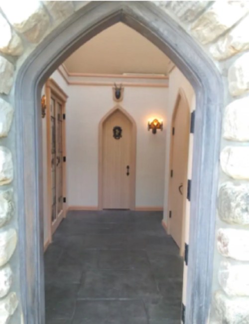 A stone archway leads into a hallway with wooden doors and wall sconces, featuring a light-colored door at the end.