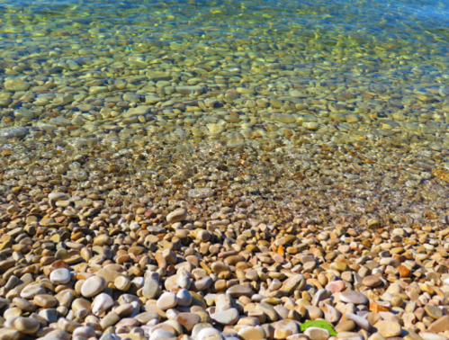 A clear, shallow water surface revealing smooth pebbles beneath, with gentle waves lapping at the shore.