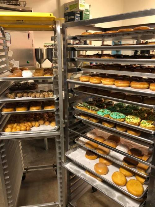 Racks filled with various types of donuts, including glazed, frosted, and decorated, in a bakery setting.