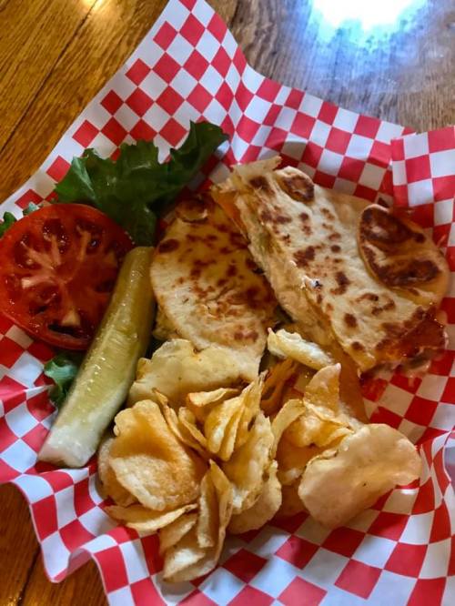 A plate with a quesadilla, potato chips, lettuce, tomato, and a pickle, served in a red and white checkered basket.
