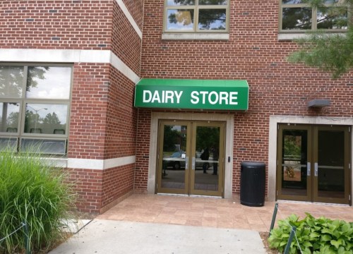 Entrance of a brick building with a green awning that reads "DAIRY STORE." Lush grass and a trash can nearby.