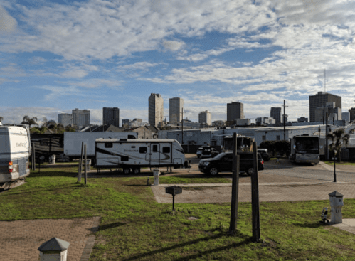 A parking area with RVs and vehicles, set against a city skyline under a partly cloudy sky.