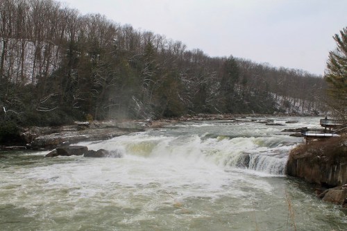 A river cascades over rocks, surrounded by trees and a snowy landscape in the background.