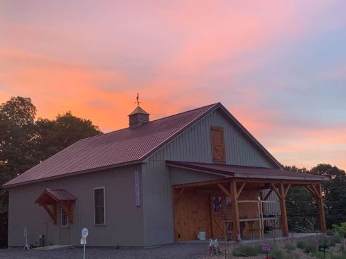A rustic barn with a metal roof under a colorful sunset sky, surrounded by trees.