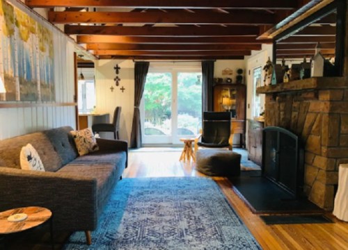 Cozy living room with a stone fireplace, wooden beams, a sofa, and large windows overlooking a garden.