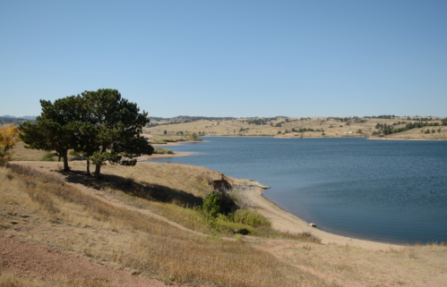 A serene lake surrounded by dry grass and hills, with a few trees along the shoreline under a clear blue sky.
