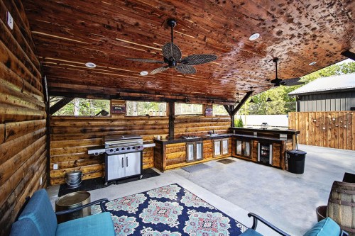 Outdoor kitchen with wooden walls, a grill, sink, and seating area, featuring a patterned rug and ceiling fans.