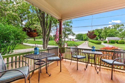 A cozy porch with two tables, chairs, hanging plants, and American flags, overlooking a green landscape and driveway.