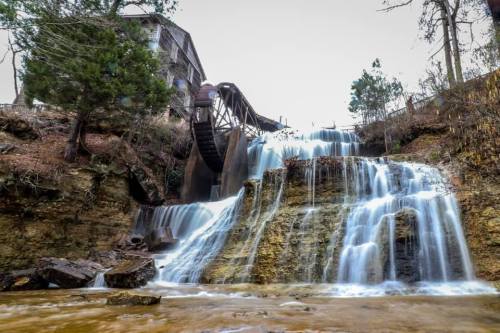 A scenic waterfall cascading over rocky terrain, with an old mill structure in the background surrounded by trees.