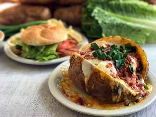 A loaded baked potato topped with cheese, bacon, and green onions, alongside a fresh salad and sandwich in the background.