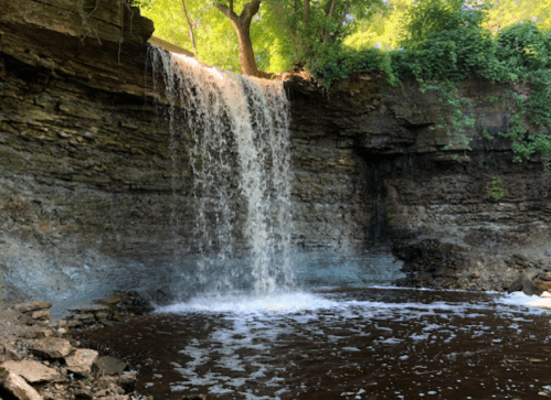 A serene waterfall cascading over rocky cliffs into a dark pool, surrounded by lush greenery and sunlight.