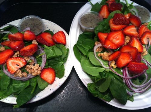 Two plates of fresh spinach salad topped with sliced strawberries, red onion, and a small cup of dressing on the side.