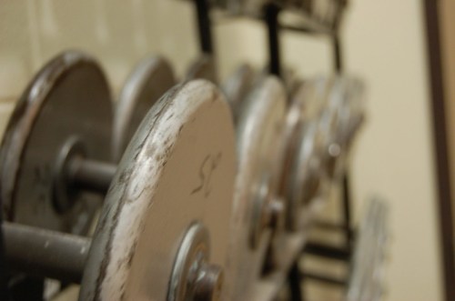A close-up of a rack of metal weight plates, slightly out of focus, with a blurred background.