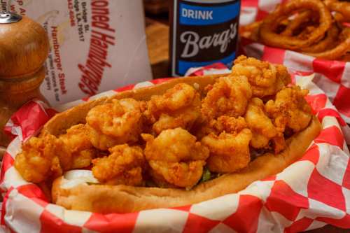 A close-up of a shrimp po' boy sandwich with crispy shrimp on a bun, served with a drink and onion rings in the background.