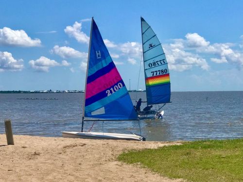 Two colorful sailboats on a sandy beach, with a calm water backdrop and fluffy clouds in a blue sky.