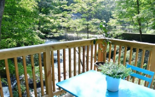 A wooden deck with a blue table and potted plant overlooks a serene, green landscape and a calm water body.