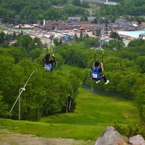 Two people ziplining down a green hillside, with a resort and water park visible in the background.