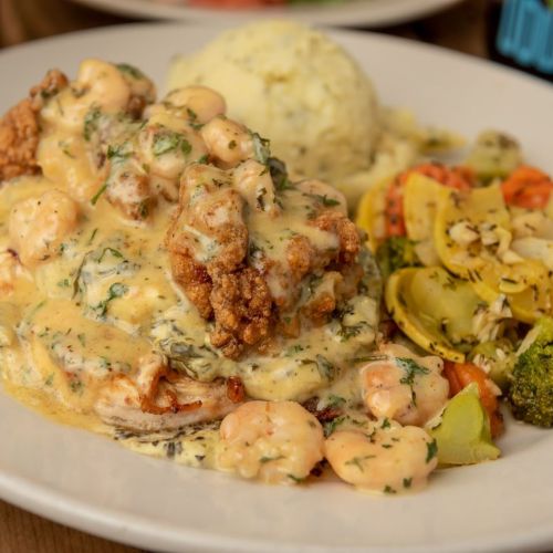 A plate of fried chicken topped with shrimp and creamy sauce, served with mashed potatoes and mixed vegetables.