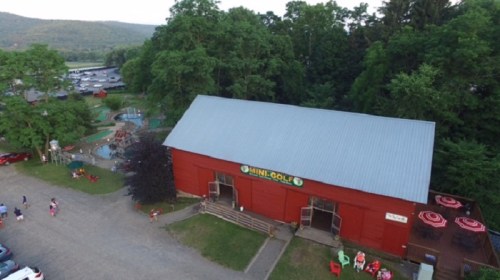 Aerial view of a red barn-style mini-golf facility surrounded by trees and outdoor seating.